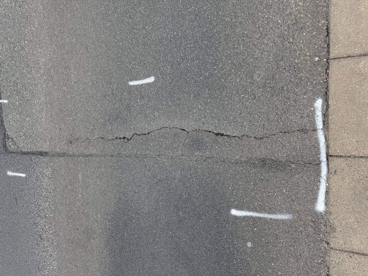 Ein durchgehender Riss im Asphalt der Fahrbahn, der entlang der Pflasterrinne verläuft und mit weißen Vermessungsmarkierungen gekennzeichnet ist.