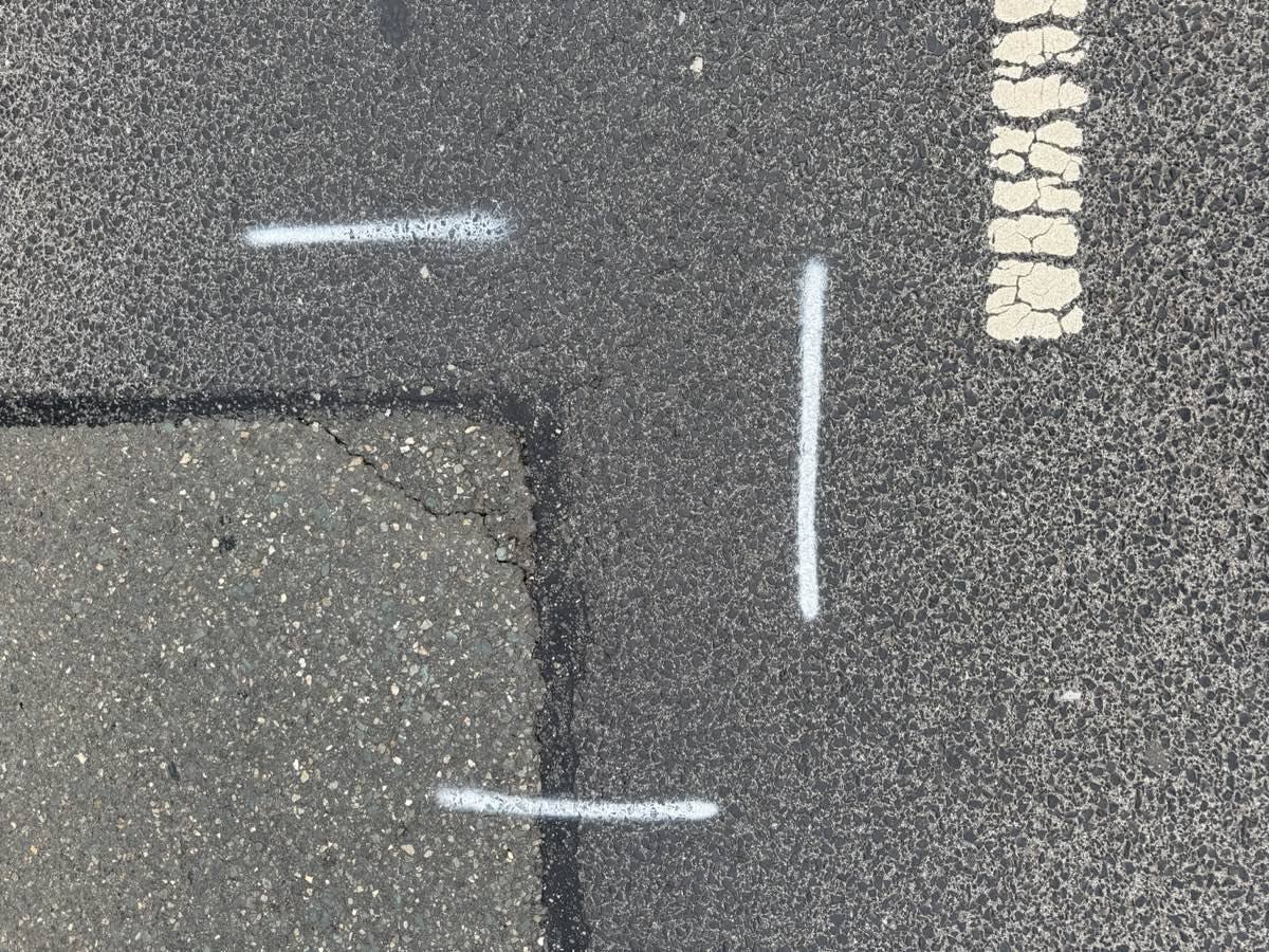 Beschädigte Fahrbahn unserer Straße: ein ausgebesserter Asphaltflicken mit aufgerissenen Rändern, weiße Vermessungsmarkierungen auf dem Belag und eine rissige Fahrbahnmarkierung.